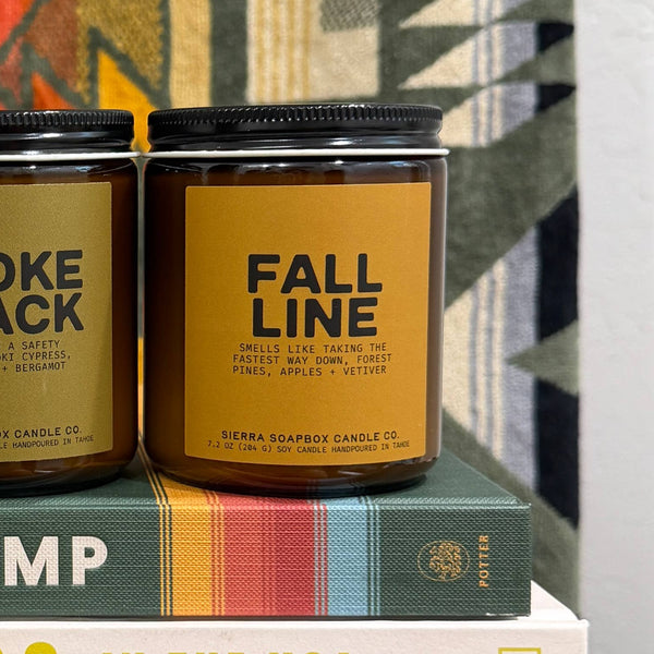 A brown glass jar candle with a golden yellow label with 'FALL LINE' and additional brand info listed below. It sits next to another candle atop a hardcover book and a blanket-covered wall.