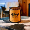 Candle labeled 'Locals Only' on a stone surface with a fireplace and armchair in the background