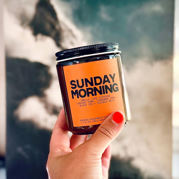 Hand holding a candle labeled 'Sunday Morning' in front of a blurred background