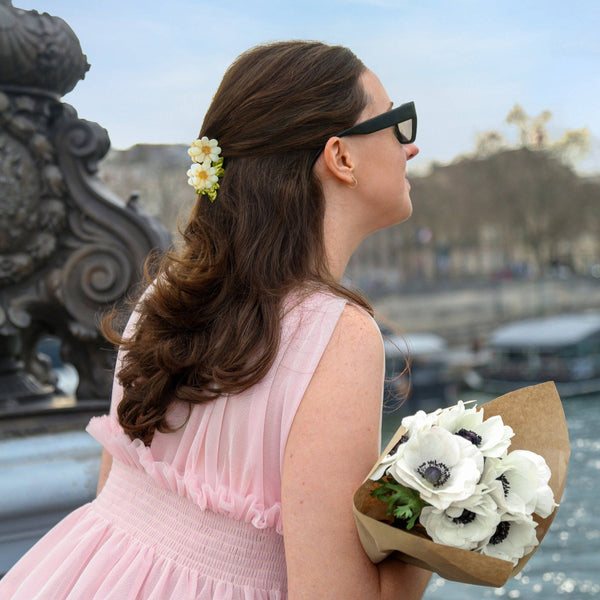 Woman in a pink dress wearing a daisy hair claw and holding flowers by a waterfront with a scenic background