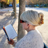 Person with blonde hair held up with a guitar shaped hair claw and sunglasses reading a book in a park with trees and benches.