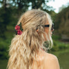 A woman with long, wavy blonde hair seen from the back, wearing a red and green hair clip and black sunglasses, with a blurred outdoor background of greenery and water.