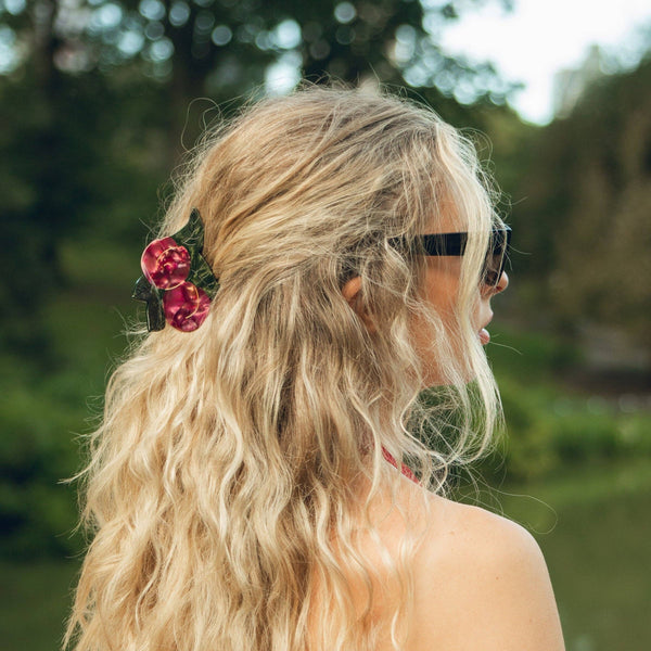 A woman with long, wavy blonde hair seen from the back, wearing a red and green hair clip and black sunglasses, with a blurred outdoor background of greenery and water.