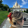Person with a red bow shaped hair clip standing on steps leading to a large white basilica under a blue sky.