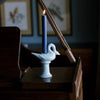 Decorative light blue swan candle holder with a dark blue candle on a wooden surface, with framed pictures in the background.