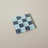 A glass mosaic tile coaster in dark teal and aqua shades on a neutral background.