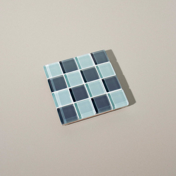 A glass mosaic tile coaster in dark teal and aqua shades on a neutral background.