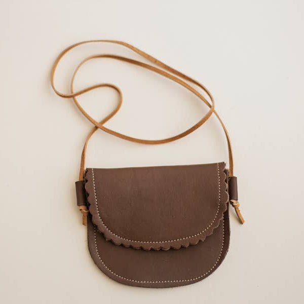 A kid-sized brown leather crossbody bag with a scalloped edge on the flap and a thin tan leather shoulder strap, photographed against a plain off-white background.