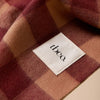 A close-up of a soft gingham scarf in muted burgundy, dusty rose, and tan tones, with a white fabric label reading 'tbco.' sewn onto one section.