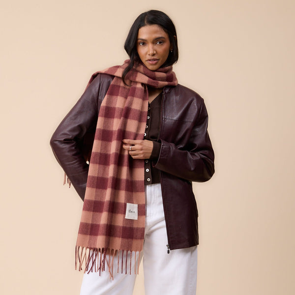 A person with dark hair wearing a dark brown leather jacket and white pants with an oversized red and tan gingham fringed scarf, standing against a neutral wall.