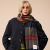 A person with blond hair wearing a dark jacket and a richly colored plaid wool scarf, standing against a neutral wall.