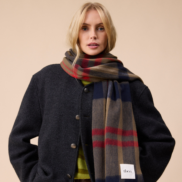 A person with blond hair wearing a dark jacket and a richly colored plaid wool scarf, standing against a neutral wall.