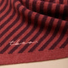 A close-up view of a berry red and chestnut striped fine knit scarf with 'tbco' embroidered logo at the edge.