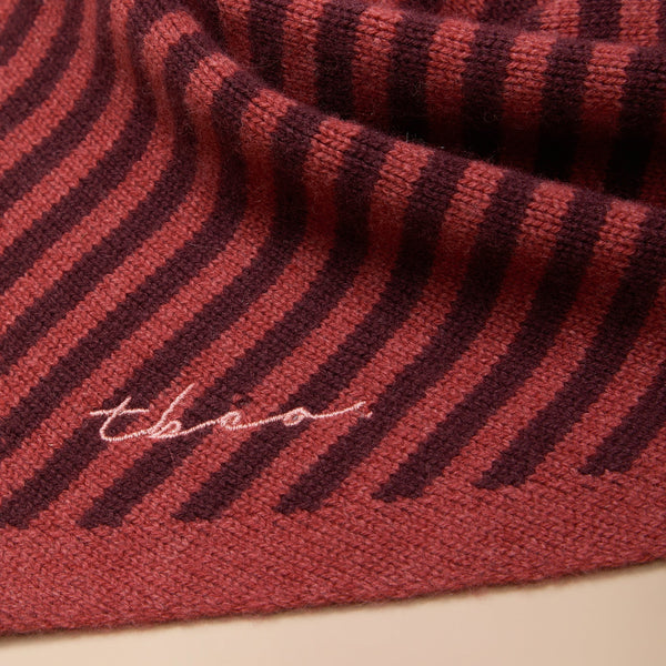 A close-up view of a berry red and chestnut striped fine knit scarf with 'tbco' embroidered logo at the edge.