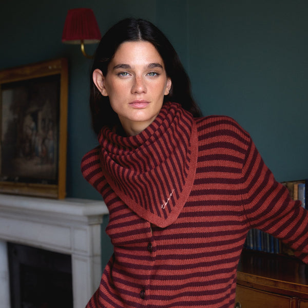 A person with long dark hair standing in a dark colored living room, wearing a berry red and chestnut striped trangle scarf and matching cardigan.