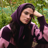 A person with long dark hair sitting outdoors wearing a rich purple triangle scarf with a purple and pink sweater.