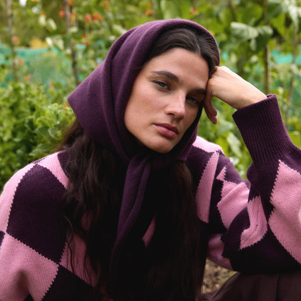 A person with long dark hair sitting outdoors wearing a rich purple triangle scarf with a purple and pink sweater.