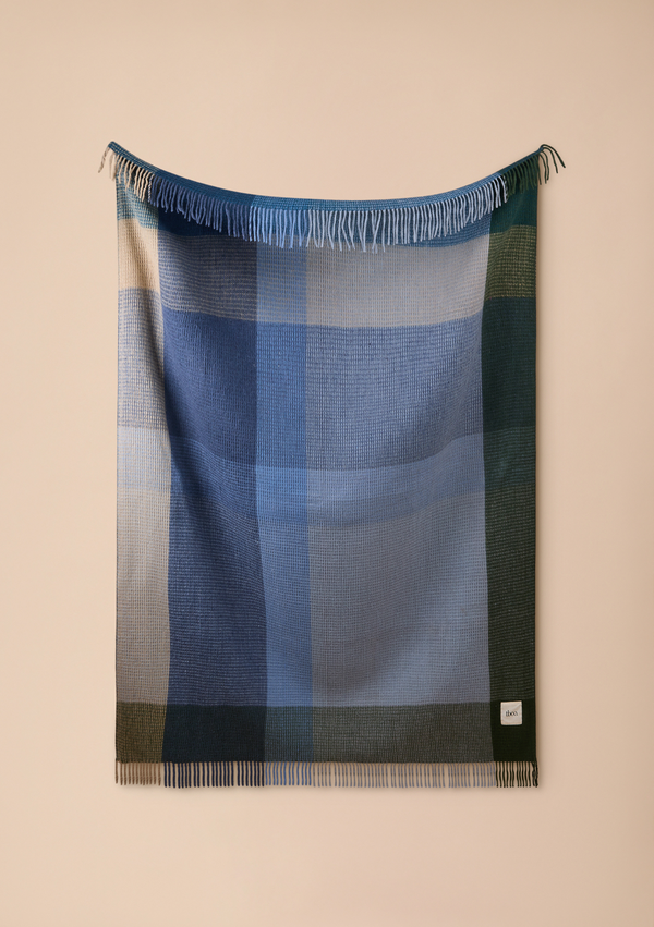 A woven plaid blanket with fringe on both ends, displayed hanging against a solid beige wall. The blanket features a color-blocked plaid pattern in shades of blue, navy, green, and beige.
