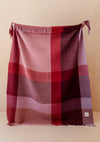 A large throw blanket with fringe on both ends featuring a large-scale plaid design in shades of burgundy, pink, red, and mauve. The blanket is being held up by hands against a light wood wall.