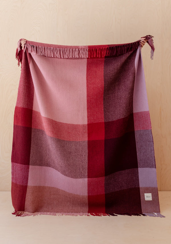A large throw blanket with fringe on both ends featuring a large-scale plaid design in shades of burgundy, pink, red, and mauve. The blanket is being held up by hands against a light wood wall.