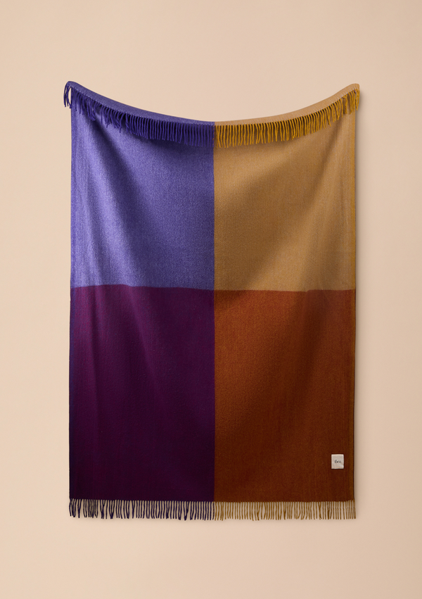 A large rectangular woven throw blanket with fringe on two edges, featuring a four-quadrant color-block pattern in purple, blue, tan, and rust brown, hanging against a solid beige wall.