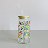 A glass travel cup with bamboo lid and glass straw. The cup is printed with a colorful array of tropical fruits. 