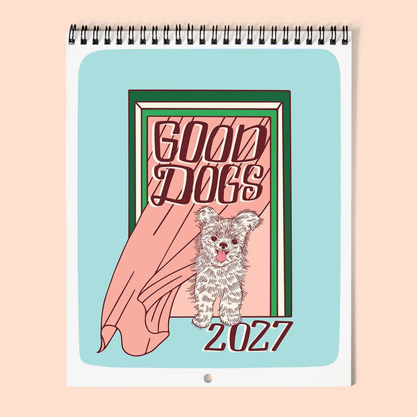 A spiral-bound 2027 wall calendar featuring a cute cartoon dog illustration and the words 'GOOD DOGS 2027' on a light blue background, shown against a soft peach backdrop.
