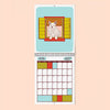A spiral-bound wall calendar page showing April 2027, with an illustrated white dog sitting in a window on a light blue background above a monthly calendar grid. The calendar is centered on a pale peach background.