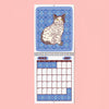 A spiral-bound April 2027 calendar page featuring a blue patterned background with an illustrated cat and a monthly grid below, shown against a solid pink background.