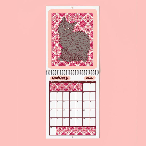 A spiral-bound wall calendar page featuring a gray cat illustration on a pink damask-patterned background, with the month October 2027 and a calendar grid below on a light pink backdrop.
