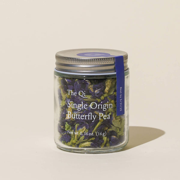 Glass jar with butterfly pea flowers labeled 'The Qi Single Origin Butterfly Pea' on a beige background