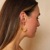 A close-up side profile of a woman wearing a large, minimalist gold crescent hoop earring. The earring features a flat, polished gold crescent shape suspended from a thin gold wire hoop that wraps around her ear.