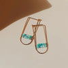 A pair of gold hoop earrings in a deep U shape with a double bar of tiny turquoise beads across the lower half laying against a shadowed beige surface. 