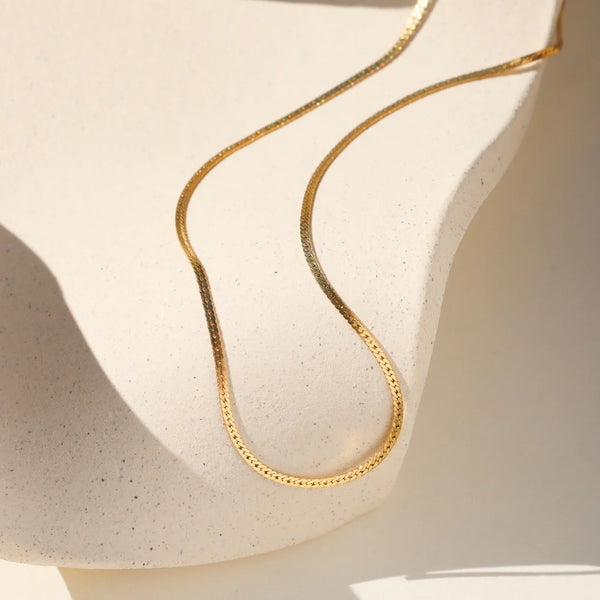 Gold herringbone chain necklace on a textured beige surface