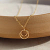 A delicate gold necklace with a double-ring pendant, resting on a curved wooden block over a light beige stone surface.