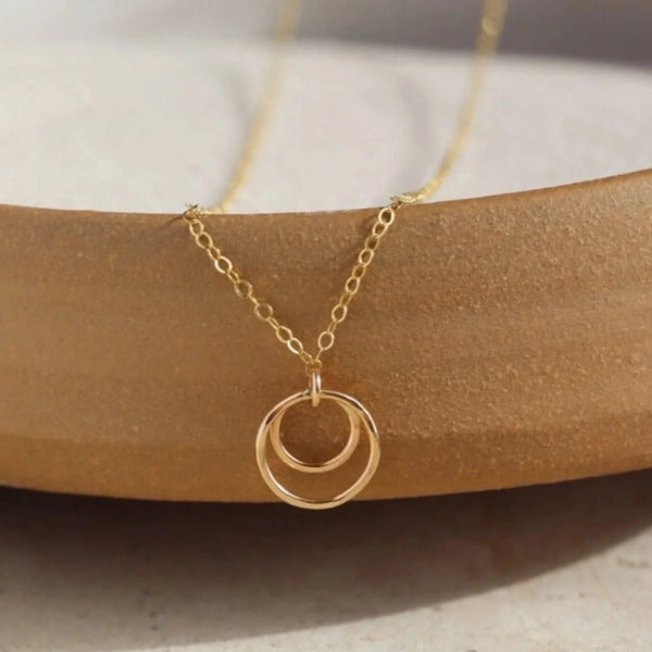 A delicate gold necklace with a double-ring pendant, resting on a curved wooden block over a light beige stone surface.