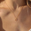 A close-up of a model's neck and chest wearing a delicate gold chain necklace with a small double-ring pendant, captured in warm natural sunlight.