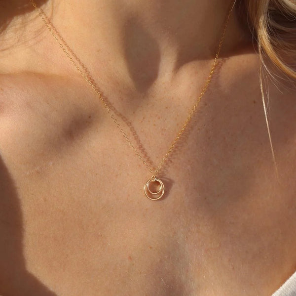 A close-up of a model's neck and chest wearing a delicate gold chain necklace with a small double-ring pendant, captured in warm natural sunlight.