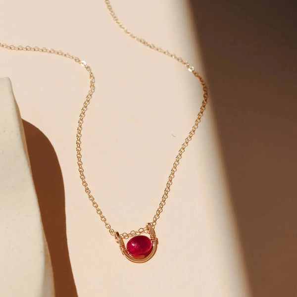 A delicate gold chain necklace with a small oval ruby gemstone and gold arc pendant, displayed on a warm beige surface with soft shadows.