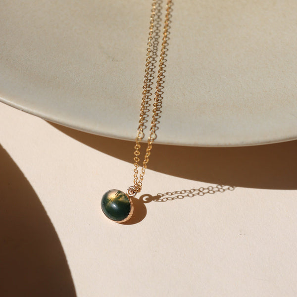 A fine gold chain necklace with an oval moss agate pendant against a shadowy neutral backdrop. 