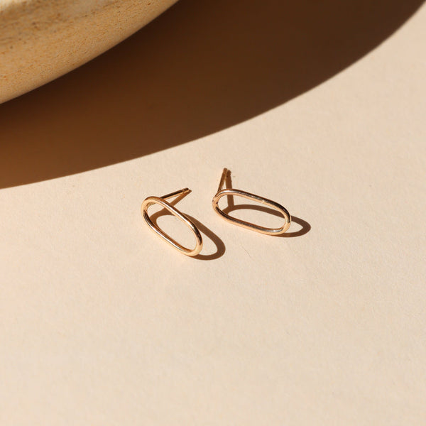 A pair of gold hollow oval shaped stud earrings against a shadowy neutral background. 