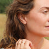 A person with brown wavy hair wearing a gold oval shaped stud earring with a blurred outdoor backdrop.