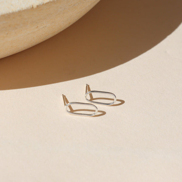 A pair of sterling silver hollow oval shaped stud earrings on a shadowy neutral background.