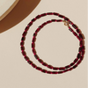 A beaded necklace made of faceted dark red garnet and rubellite beads, coiled on a warm beige background with a partial cream jewelry display stand visible in the top left.