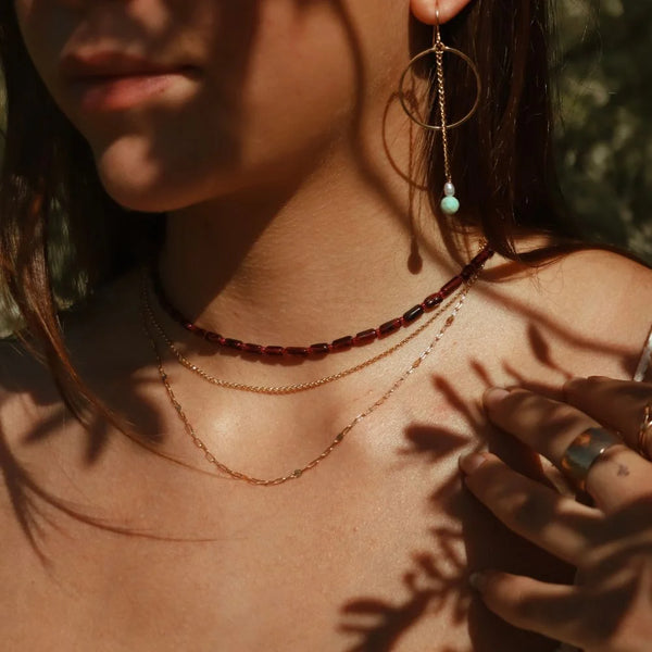 Close-up of a woman wearing layered necklaces, including a dark red beaded choker, a thin gold chain, and a delicate gold chain, with a dangling gold hoop earring featuring a light blue bead. Warm sunlight and leaf shadows are cast across her skin.