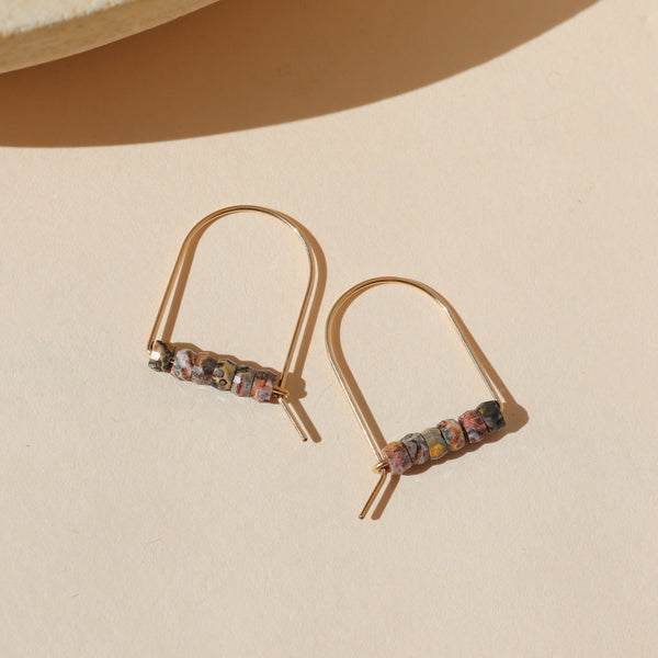 A pair of gold arch earrings with tiny leopard jasper beads strung across a bottom bar, sitting against a neutral background. 