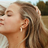 A person with dark blonde hair standing outside, wearing a silver moon sliver shaped stud earring.