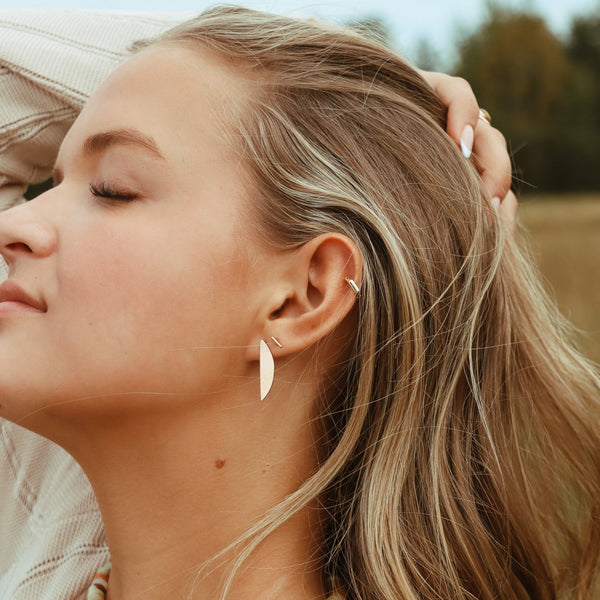 A person with dark blonde hair standing outside, wearing a silver moon sliver shaped stud earring.