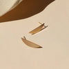 A pair of gold slim crescent shaped stud earrings on a shadowy neutral background.