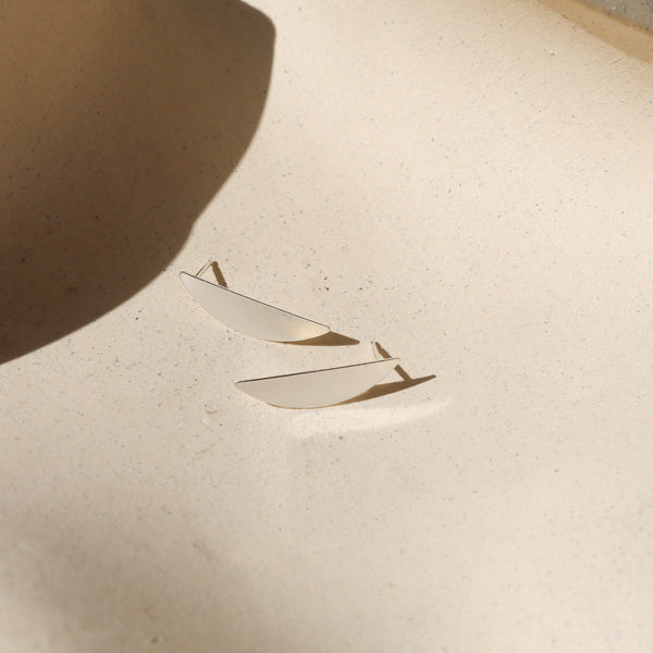 A pair of sterling silver moon sliver shaped stud earrings on a shadowy neutral background.